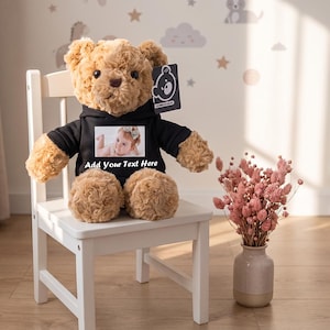 Puede incluir: Un oso de peluche marrón claro con una sudadera negra con capucha. La sudadera presenta una impresión fotográfica rectangular de un bebé y el texto blanco Add Your Text Here. Una etiqueta negra con un icono de oso blanco está en la oreja.