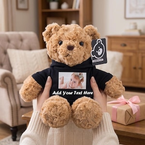 Può includere: Un orsacchiotto di peluche marrone chiaro che indossa una felpa nera con cappuccio. La felpa presenta una stampa fotografica rettangolare di un bambino e il testo bianco Add Your Text Here. Un'etichetta rettangolare nera con un'icona di orso è attaccata all'orecchio.