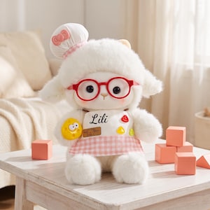 May include: A white plush lamb toy wearing a chef hat with a pink bow, red round glasses, and a white apron featuring a pink gingham trim. The apron displays the name Lili in black script, a small bear patch, and colorful fruit-shaped accents.