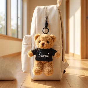 Puede incluir: Una mochila de color beige claro con un llavero de osito de peluche de color marrón claro sujeto a la cremallera. El osito lleva una camiseta negra con el nombre David impreso en letras blancas. Tiene ojos oscuros y nariz marrón.