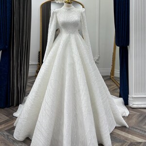 May include: A floor-length white gown featuring a high neckline, long sleeves with puffed shoulders, and a full, voluminous skirt. The entire garment is covered in intricate, shimmering beadwork and sequins arranged in a geometric pattern.
