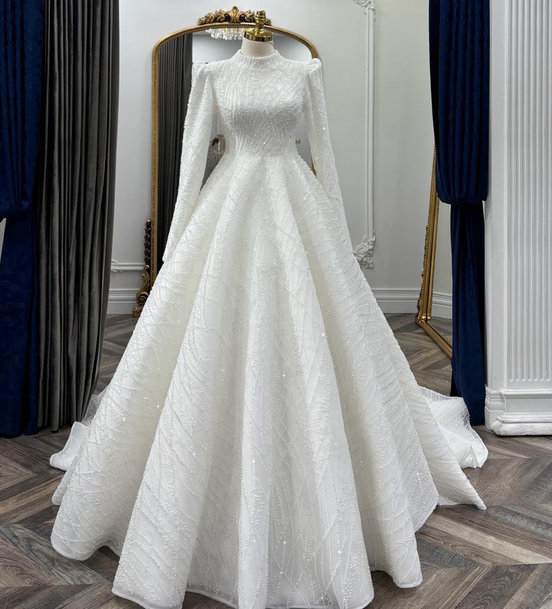 May include: A floor-length white gown featuring a high neckline, long sleeves with puffed shoulders, and a full, voluminous skirt. The entire garment is covered in intricate, shimmering beadwork and sequins arranged in a geometric pattern.