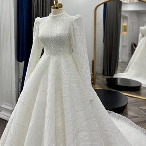 May include: A floor-length white gown on a mannequin featuring a high neckline, long sleeves with puffed shoulders, and a full skirt. The entire garment is covered in intricate white beadwork and sequins arranged in a swirling, geometric pattern.