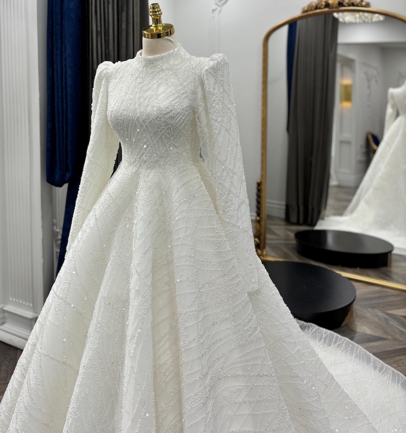 May include: A floor-length white gown on a mannequin featuring a high neckline, long sleeves with puffed shoulders, and a full skirt. The entire garment is covered in intricate white beadwork and sequins arranged in a swirling, geometric pattern.