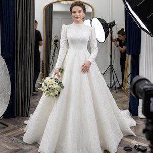 May include: A floor-length white wedding gown with long sleeves, a high neckline, and a full, voluminous skirt. The fabric features a subtle, shimmering textured pattern throughout. The garment is paired with a bouquet of white calla lilies.