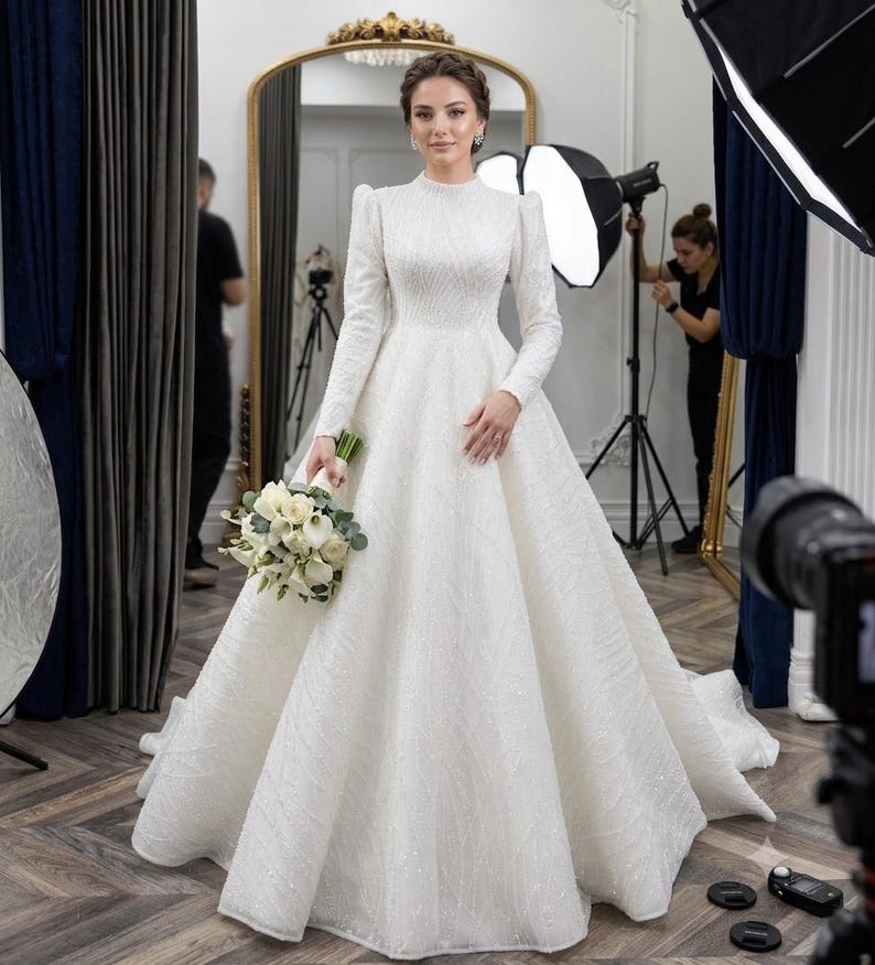 May include: A floor-length white wedding gown with long sleeves, a high neckline, and a full, voluminous skirt. The fabric features a subtle, shimmering textured pattern throughout. The garment is paired with a bouquet of white calla lilies.