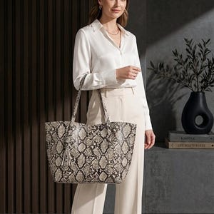 May include: A large tote bag with a snakeskin pattern in shades of grey, brown, and cream. The bag has long handles and a tassel. The bag is held by a person wearing a white blouse and cream-coloured trousers.