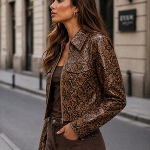 Peut inclure: Veste marron et noire &agrave; motif peau de serpent, avec un col et une fermeture &eacute;clair. La veste a deux poches avant et est port&eacute;e avec un pantalon marron et un haut marron. La tenue est port&eacute;e &agrave; l'ext&eacute;rieur.