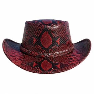 Puede incluir: Un sombrero de vaquero con un estampado de piel de serpiente en rojo y negro. El sombrero tiene una banda trenzada alrededor de la corona y un ala ancha. El sombrero es de cuero.