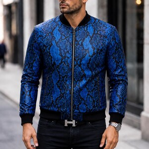 Puede incluir: Chaqueta bomber de cuero azul con estampado de piel de serpiente, con cuello, puños y dobladillo negros. La chaqueta tiene una cremallera plateada y se usa con jeans negros y una hebilla plateada. La persona también usa gafas de sol y un reloj.