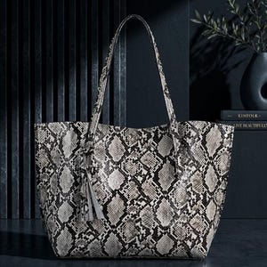 Könnte beinhalten: Große Tragetasche mit Schlangenmuster in Grau- und Weißtönen. Die Tasche hat lange Henkel und eine dekorative Quaste. Die Tasche ist aus strapazierfähigem Material gefertigt und für den täglichen Gebrauch geeignet.