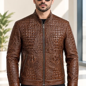 May include: A brown leather jacket with a textured crocodile skin pattern, a stand-up collar, and a full-length front zipper. The jacket features two zippered side pockets and long sleeves with cuffs. The text ALVERA leather is visible.
