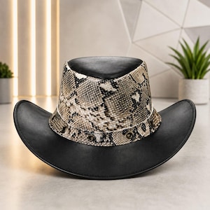 May include: A hat featuring a solid black crown and wide brim, accented by a band with a tan and brown snakeskin pattern. The band includes four metallic eyelets on the front, providing a distinct contrast between the dark and textured elements.