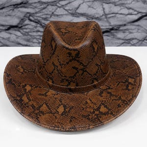 Puede incluir: Un sombrero fedora de color marrón con un patrón de piel de serpiente marrón oscuro en toda la superficie. El sombrero tiene una copa estructurada con un pliegue central y un ala ancha y curva. El material tiene una textura uniforme.