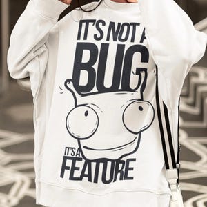 May include: White sweatshirt with the text "IT'S NOT A BUG IT'S A FEATURE" in black. The graphic features a cartoon-style alien face with large eyes and a smiling mouth. The sweatshirt has long sleeves and a crew neck.