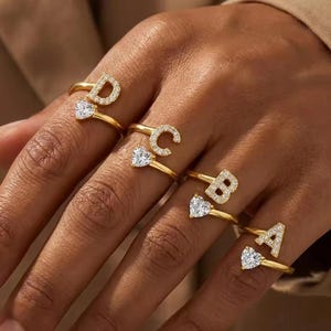 Può includere: Anelli dorati con le iniziali D, C, B e A, ciascuno ornato di pietre chiare e scintillanti. Gli anelli presentano una gemma a forma di cuore sotto l'iniziale. Gli anelli sono mostrati su una mano.