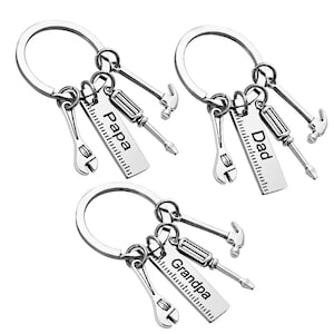 May include: Three silver-toned metal keychains, each featuring a circular ring attached to four charms: a miniature wrench, a hammer, a screwdriver, and a rectangular tag shaped like a ruler. The tags are engraved with the words Papa, Dad, and Grandpa.