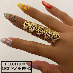 May include: Six gold-colored initial rings with ornate floral designs are displayed on a hand. The rings are in a row, each featuring a different letter. The nails are painted with various designs. The text "FREE GIFT BOX NEXT DAY SHIPPING" is in a black rectangle.