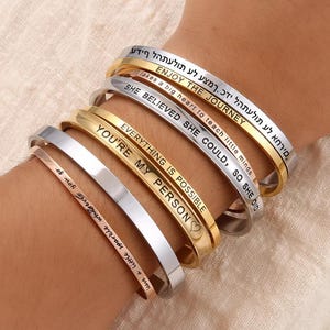 Puede incluir: Colección de seis pulseras metálicas en oro, plata y oro rosa. Cada pulsera presenta una cita inspiradora grabada. Las citas incluyen "Enjoy the Journey" y "Everything is Possible."