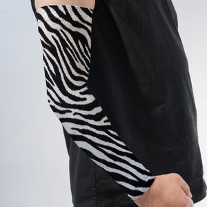 May include: An arm sleeve featuring a high-contrast black and white zebra stripe pattern. The sleeve covers the forearm and upper arm, extending from the wrist to the bicep area, and is worn over a plain black short-sleeved t-shirt.