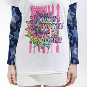 May include: A white short-sleeve t-shirt featuring a tie-dye sunflower graphic with the text DON'T WORRY BE HIPPIE and a peace sign. The shirt has long sleeves underneath with a dark blue denim-look fabric printed with light blue stars.