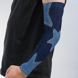 May include: An arm sleeve featuring a dark blue denim-like fabric pattern with light blue star shapes printed across the surface. The stars have a visible stitched outline design, creating a textured appearance on the fitted fabric sleeve.