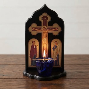 Puede incluir: Un soporte de icono de madera negra en forma de arco con un crucifijo central con las letras I.N.R.I. y dos figuras laterales. Un pequeño portavelas votivo de cristal azul profundo con una llama encendida descansa sobre la base.