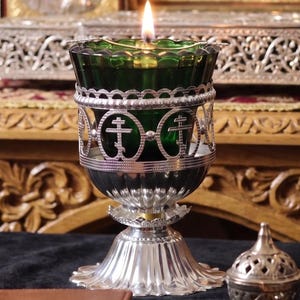 May include: A green glass oil lamp with a silver-toned metal holder featuring cut-out Orthodox cross symbols. The lamp has a fluted base and a lit wick. Beside it sits a small, ornate silver-toned metal incense burner with a perforated dome lid.