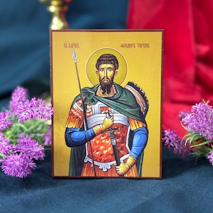 Op de afbeelding: Een rechthoekig houten icoon met een bebaarde religieuze figuur, gekleed in een harnas en een groene mantel, die een speer en een zwaard vasthoudt. De figuur staat tegen een gouden achtergrond met cyrillische tekst "СЕ БАРИКА" en "ѲЕОДОРЪ ТИРОНЪ".