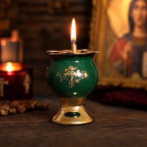 Op de afbeelding: Een groene olielamp met een goudkleurige voet en rand, voorzien van een goudkleurig kruis en bloemmotief aan de voorzijde. Een enkele vlam brandt in het midden van de lamp, die een ronde vorm heeft en een uitlopende bovenrand met inkepingen.