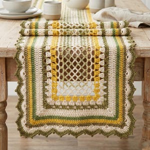 May include: A crocheted table runner with a square, open-weave center and a scalloped edge. The runner features stripes of cream, yellow, and green. Rolled up, the runner reveals the same color pattern. The runner is displayed on a wooden table.