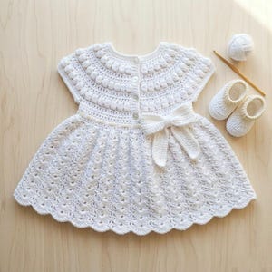 Puede incluir: Vestido de bebé blanco de ganchillo hecho a mano con lazo, mangas cortas y dobladillo festoneado. El vestido va acompañado de patucos de ganchillo a juego, un ovillo de lana y un ganchillo. El vestido mide aproximadamente 30 cm de largo.