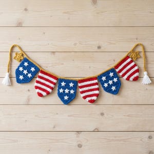 May include: A crocheted patriotic banner featuring alternating blue pennants with white stars and red and white striped pennants. The banner is connected by a gold cord and has gold star accents and white tassels.