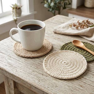 Puede incluir: Una taza de cerámica blanca llena de café oscuro se encuentra sobre un posavasos beige tejido a ganchillo. También se ven dos posavasos tejidos a ganchillo adicionales, uno crema y otro verde. Una cuchara de madera descansa sobre el posavasos verde.