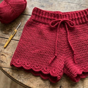 May include: A pair of handmade, red crochet shorts with a scalloped edge and drawstring waist. A ball of red yarn and a crochet hook are visible on a wooden surface, suggesting a crafting theme.