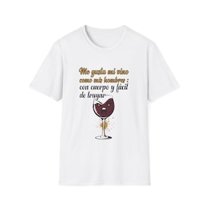 May include: White t-shirt featuring a wine glass design with a face and the Spanish text "Me gusta mi vino como mis hombres: con cuerpo y fácil de tragar" in gold lettering. The wine glass has a splash of gold and brown paint.