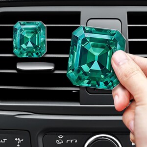 May include: Two emerald green car air fresheners shaped like faceted gemstones. The air fresheners feature a deep green color with geometric, multi-faceted surfaces that mimic the appearance of cut crystals, designed to attach to vehicle air vents.