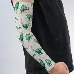May include: An arm sleeve featuring a repeating pattern of various green dinosaurs on a light gray background with small dark gray spots. The sleeve is worn on an arm, extending from the wrist to the upper arm, paired with a plain black t-shirt.