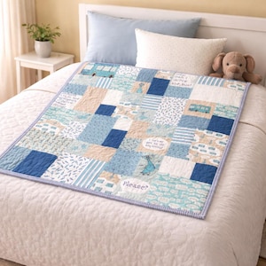 May include: A patchwork quilt in shades of blue, white, and tan, featuring various patterns and text. The quilt has a blue border and is placed on a white bed. A stuffed animal dog is on the bed.