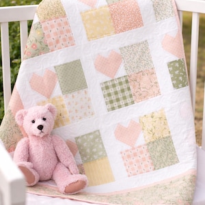 May include: A pastel-colored baby quilt with a patchwork design featuring squares of various floral and gingham patterns, and heart shapes. A pink teddy bear sits on the quilt, which is draped over a white crib.