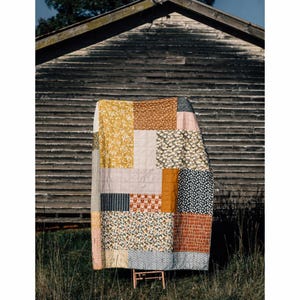 May include: A patchwork quilt with a variety of floral, geometric, and solid fabric squares in shades of yellow, brown, pink, and navy blue. The quilt is draped over a small wooden stand in front of a weathered wooden wall.