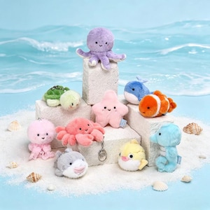 May include: A selection of plush sea creatures, including an octopus, starfish, turtle, whale, clownfish, crab, shark, pufferfish, and dolphin. The toys are in pastel colours and are arranged on a sandy surface with a blue ocean backdrop.
