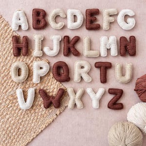 May include: A collection of crocheted alphabet letters in various colors, including white, beige, and brown. The letters are arranged in rows, showcasing the full alphabet. Yarn balls are visible in the background, suggesting a craft project.