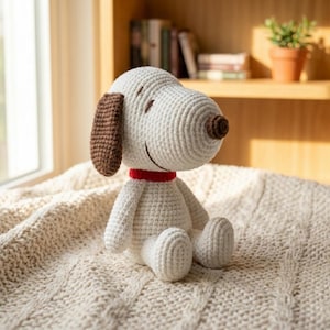 May include: A crocheted Snoopy plush toy, featuring a white body, brown ears and nose, and a red collar. The handmade Snoopy is sitting on a cream-colored blanket. The background includes a window and a wooden shelf with books and a potted plant.