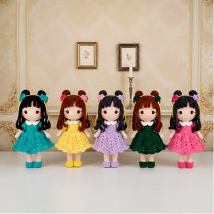May include: Five handmade crochet dolls standing in a row, each wearing a different colored dress in teal, yellow, lavender, dark green, and pink. Each doll features dark or brown hair styled with small floral hair accessories and matching shoes.