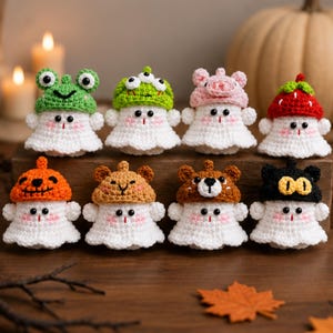 May include: Eight small, white crocheted ghost figurines arranged in two rows. Each ghost wears a different colorful crocheted hat, including designs resembling a frog, a mushroom, a pig, a strawberry, a pumpkin, a bear, and a black cat.