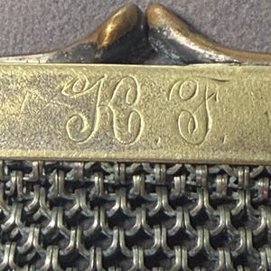 May include: Close-up of an antique metal mesh purse. The purse features a brass frame with the initials "KC F" engraved on it. The mesh is made of small, interlocking metal rings, creating a textured surface. The clasp is a curved, bronze-colored metal.
