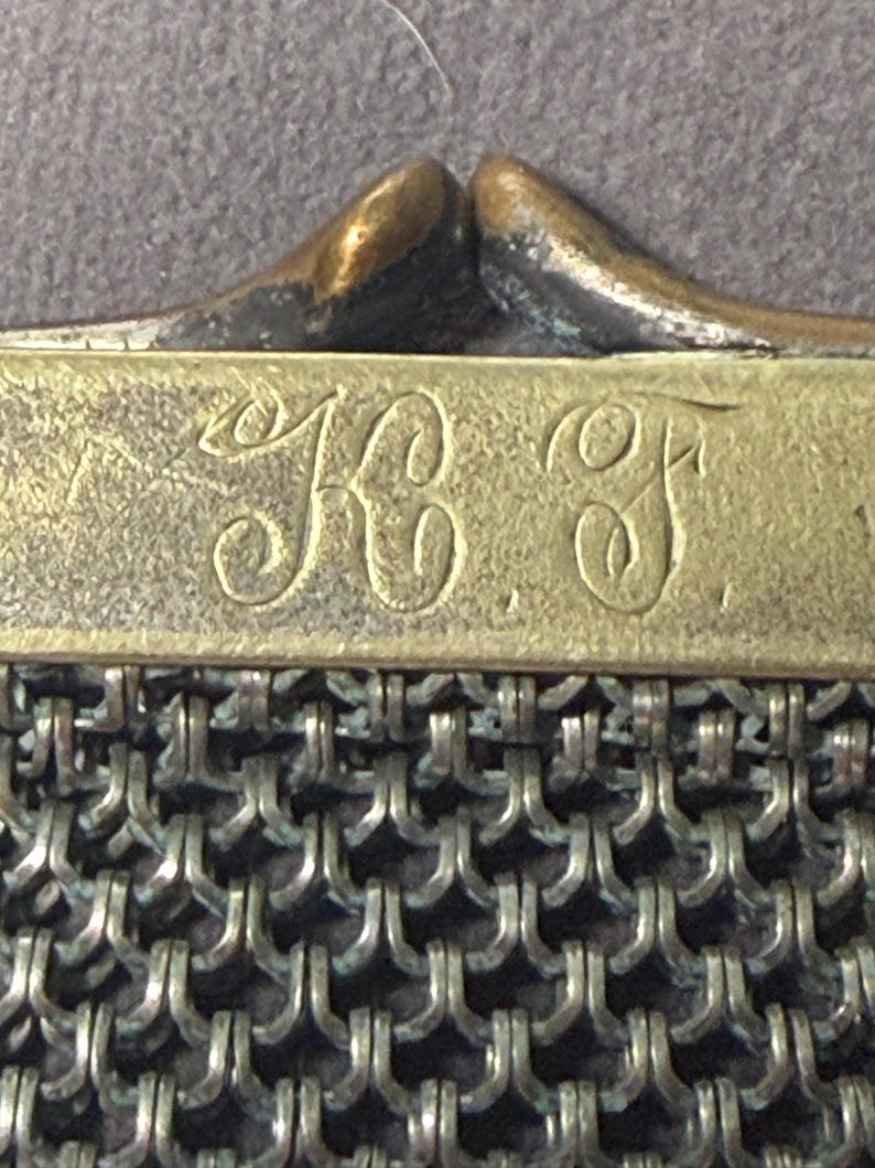 May include: Close-up of an antique metal mesh purse. The purse features a brass frame with the initials "KC F" engraved on it. The mesh is made of small, interlocking metal rings, creating a textured surface. The clasp is a curved, bronze-colored metal.