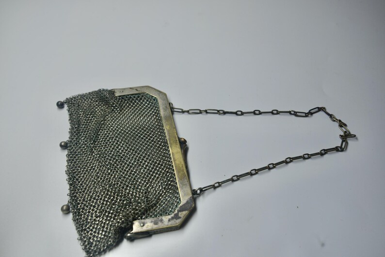 May include: Antique silver mesh purse with a chain handle. The purse has a rectangular frame and a delicate, woven mesh body. Small silver beads adorn the bottom edge. The chain handle is also silver.