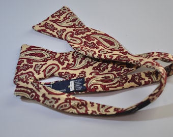 May include: A paisley patterned bow tie in shades of cream and burgundy. The bow tie has a black adjustable strap.